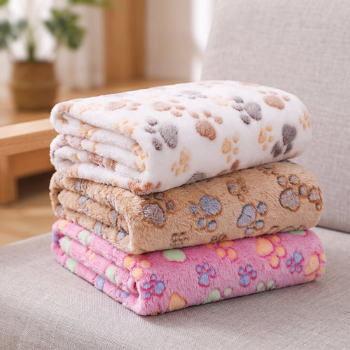 Stack of three paw print fleece pet blankets in white, tan and pink on a sofa cushion
